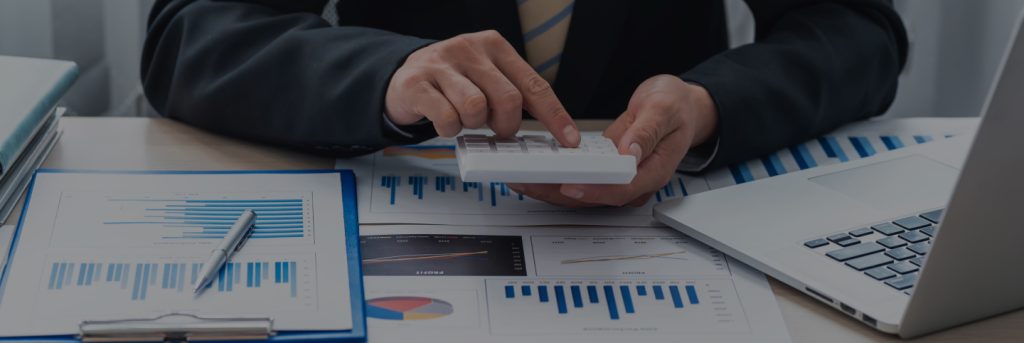 Uomo in giacca e cravatta che utilizza una calcolatrice su una scrivania con laptop e documenti contenenti grafici e report finanziari